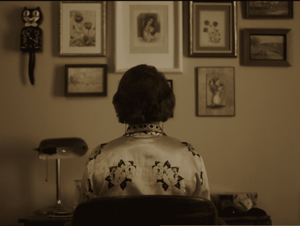 Claude is in front of her computer, there are frames all over her wall, she is wearing a flowered blouse